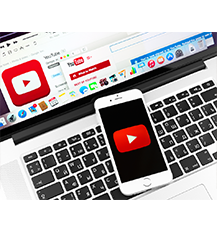 Graphic of a silver computer with black keys which has a phone placed on it with the logo for YouTube which is a red square with a white play button in it. On the screen of the computer there are many applications on the bottom bar and YouTube is open on the computer.