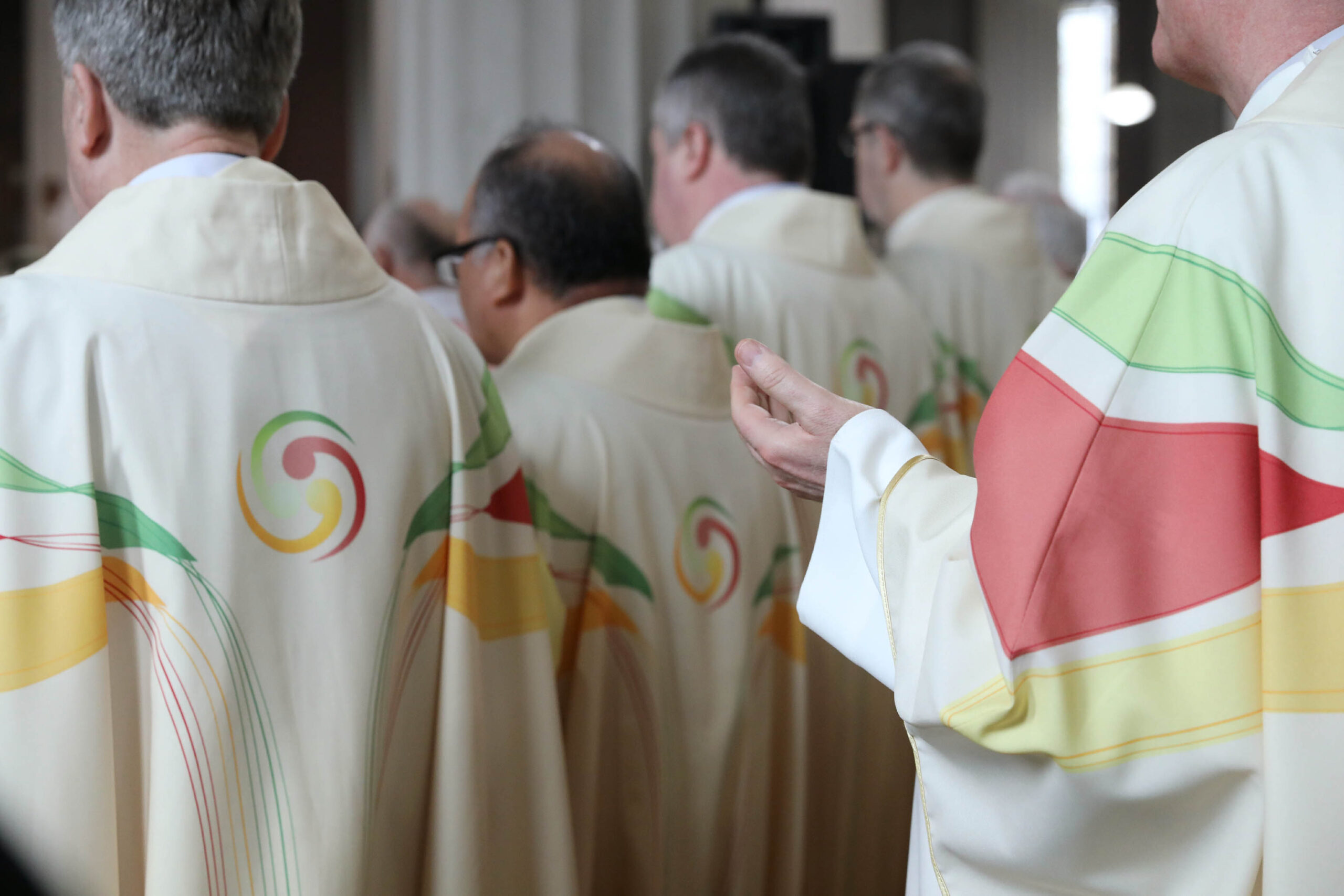 Graphic of a group of priests facing towards the congregation in white robes with red green and yellow stripes and a swirl of those colours on their backs. One priest has a hand outstretched.