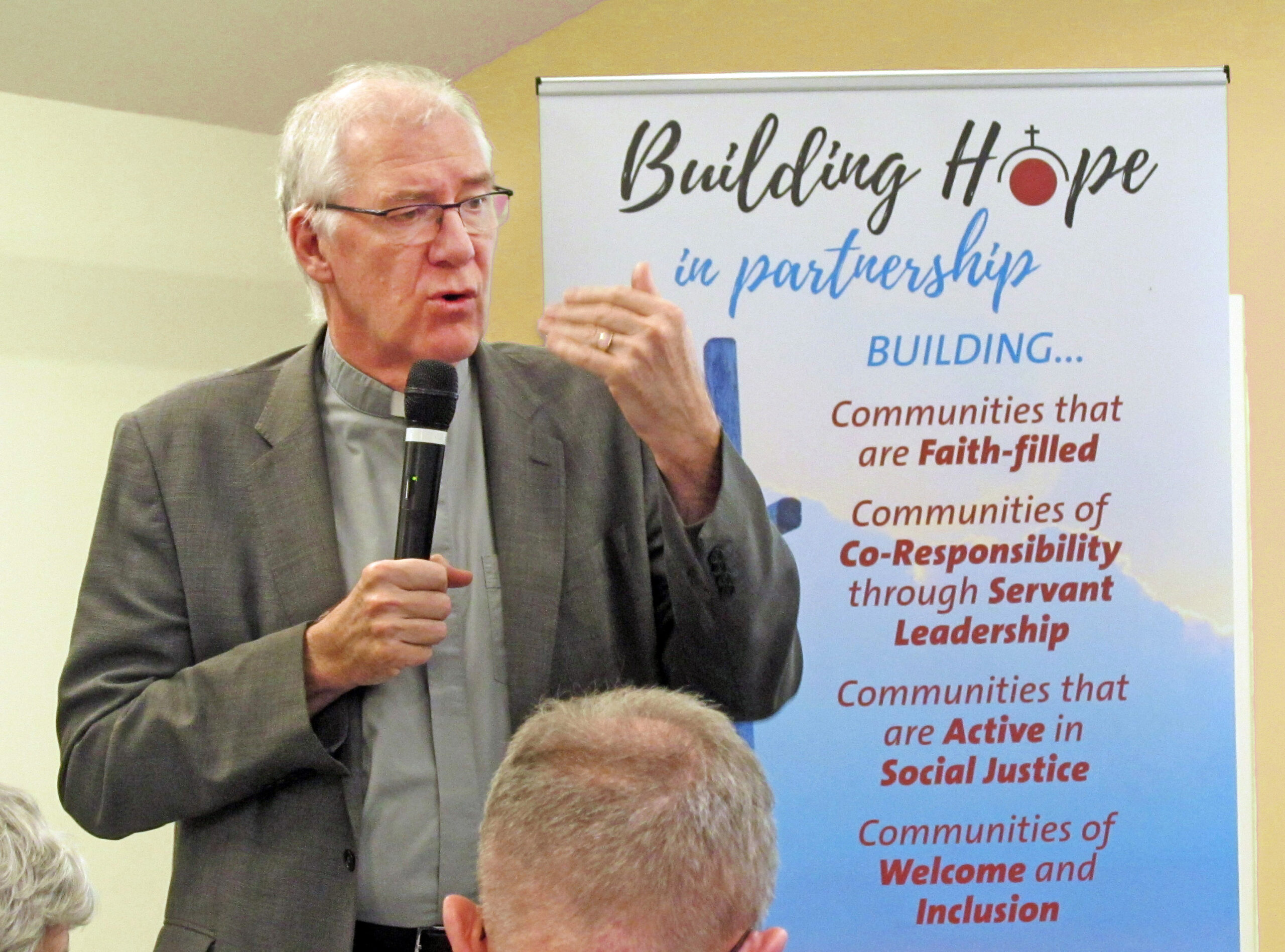 Graphic of a man with a clerical collar speaking into a microphone to a group of people. Behind him is a large banner for Building Hope in partnership.