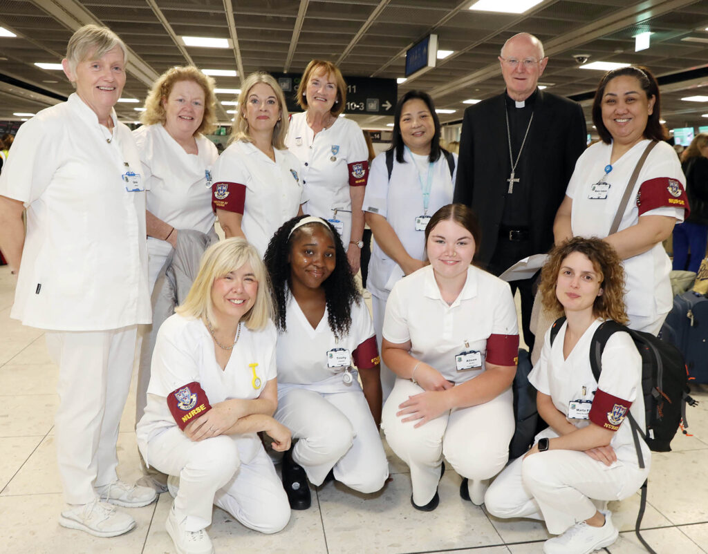 Graphic for the Dublin Diocesan Pilgrimage to Lourdes 2025. Picture of Archbishop Farrell with some of the nurses at Dublin Airport. The group are all smiling at the camera with the nurses wearing white nursing suits and Archbishop Farrell wearing a black priest suit and clerical collar with a cross around his neck.