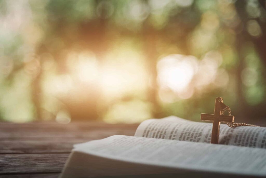 Graphic depicts a Bible with a cross in the middle on a wooden table with trees in the background and a golden sun.